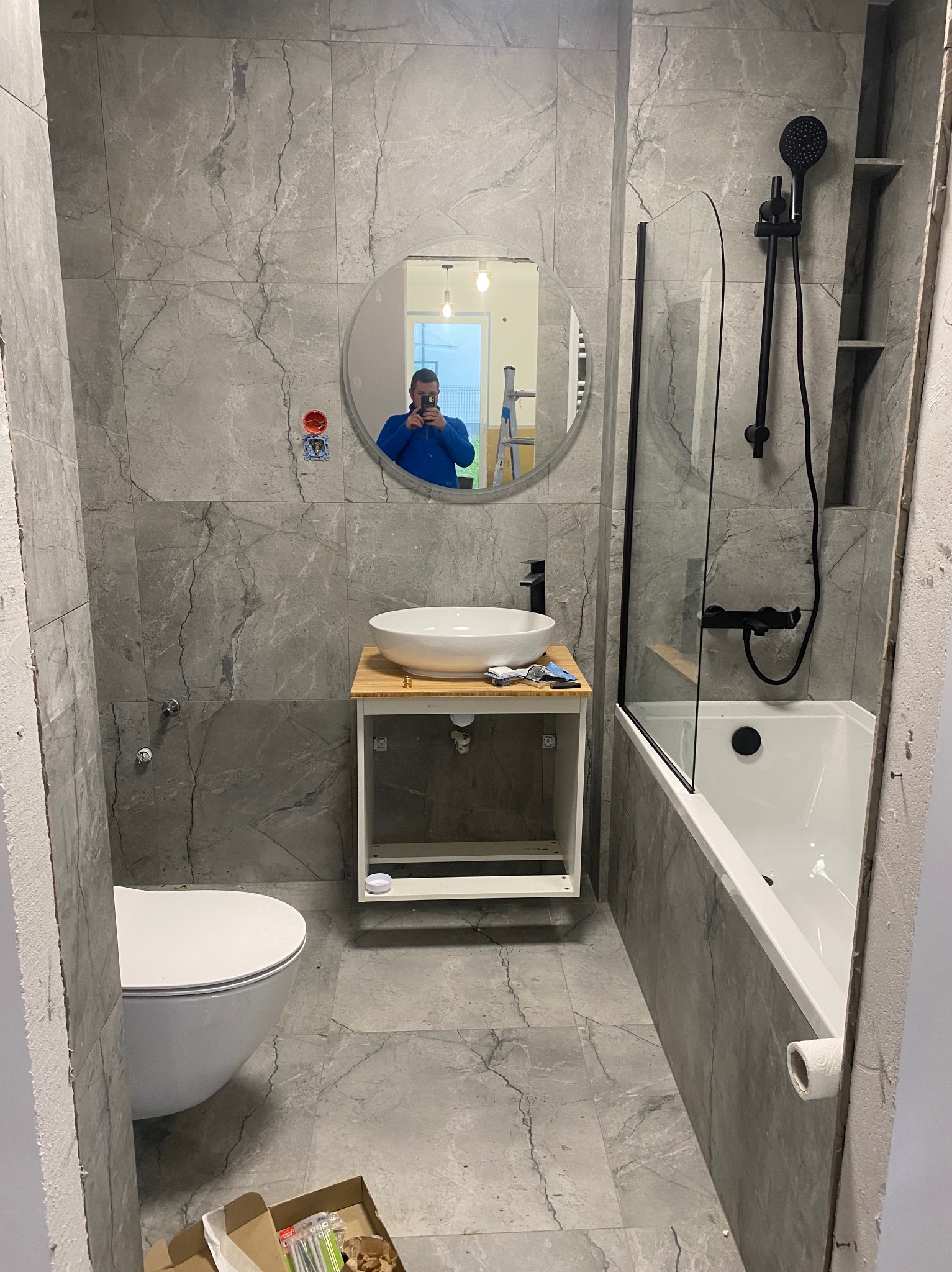 Łazienka w trakcie prac wykończeniowych: szare płytki imitujące marmur na ścianach i podłodze, biała umywalka na drewnianym blacie, toaleta wisząca, wanna z czarną armaturą i szklanym parawanem...