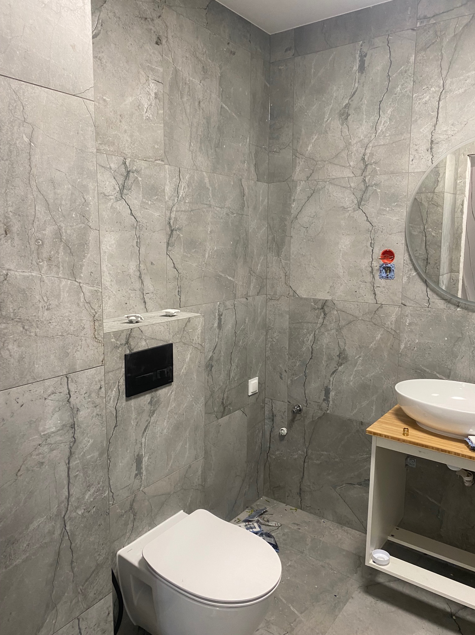 Wykończona łazienka w szarościach z marmurkowymi płytkami, wisząca toaleta z czarnym przyciskiem, umywalka nablatowa na szafce z bambusowym blatem, okrągłe lustro, widoczne niedociągnięcia...