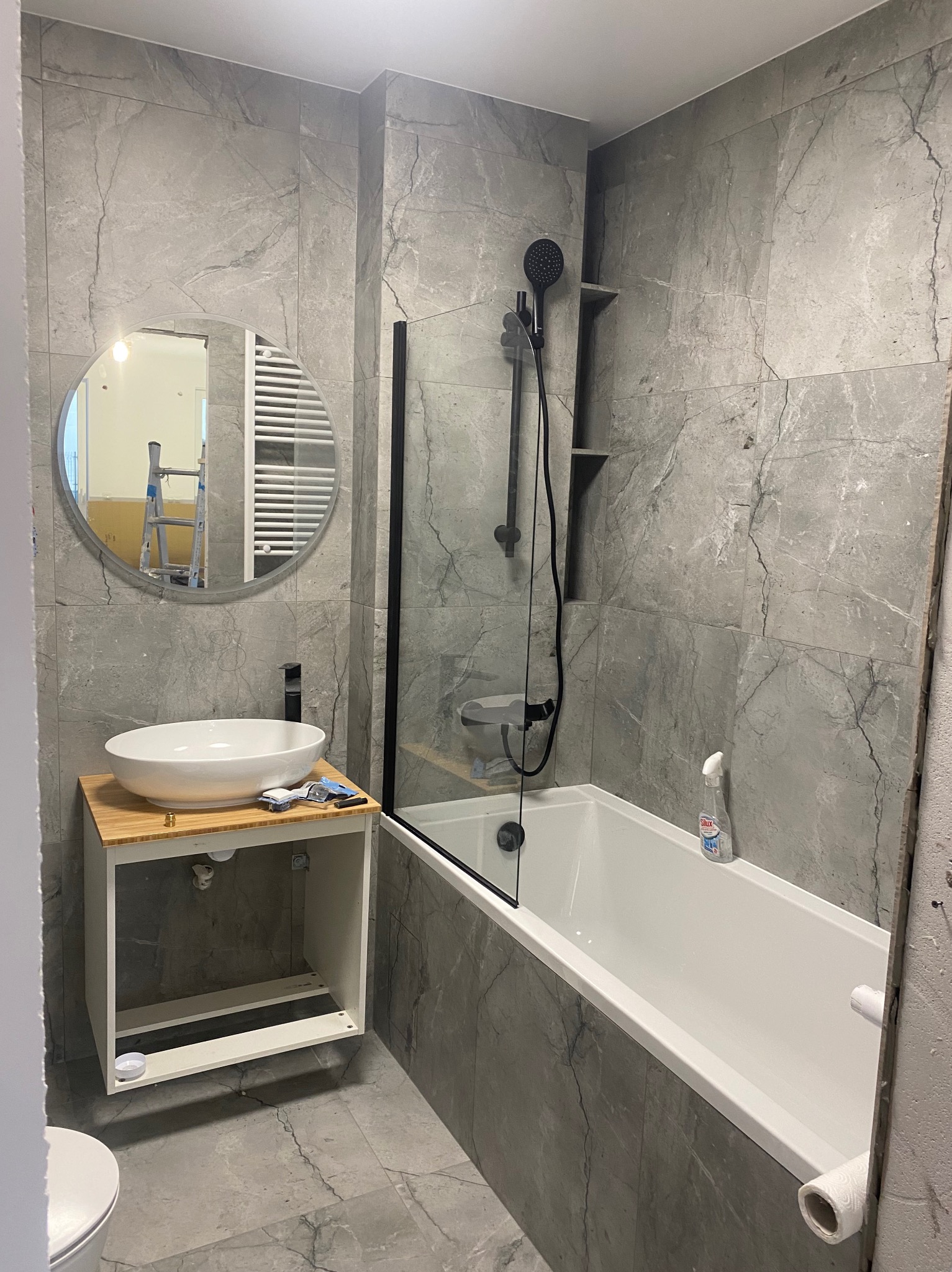 Łazienka w trakcie remontu: szare płytki imitujące marmur, biała wanna z czarną armaturą, umywalka nablatowa na szafce z bambusowym blatem, okrągłe lustro z widoczną drabiną w odbiciu.