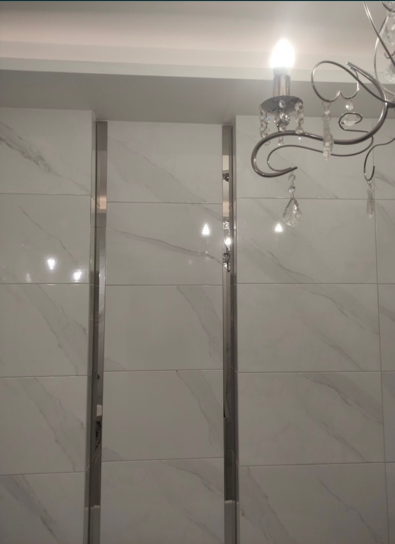 Białe płytki imitujące marmur na ścianie, oddzielone pionowymi, metalowymi listwami; fragment ozdobnego żyrandola z kryształkami w prawym górnym rogu.