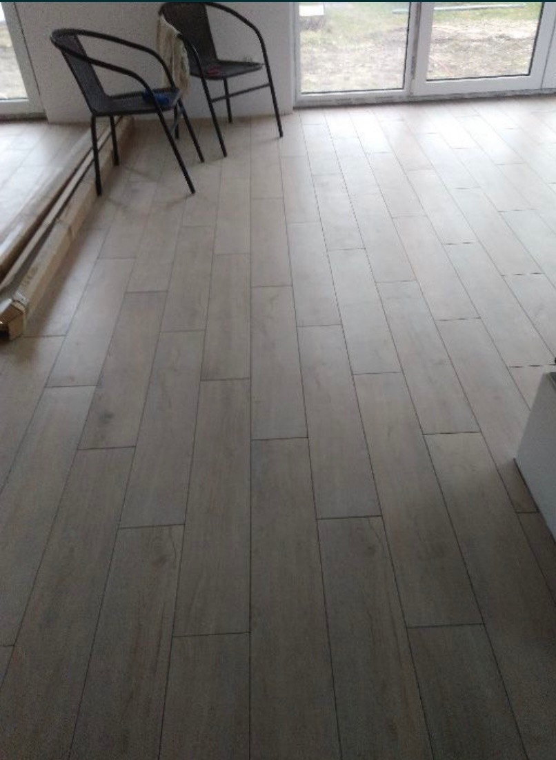 Ułożona podłoga z paneli w odcieniach szarości, widoczne dwa czarne krzesła i paczka paneli w rogu pomieszczenia, światło dzienne wpada przez okno.