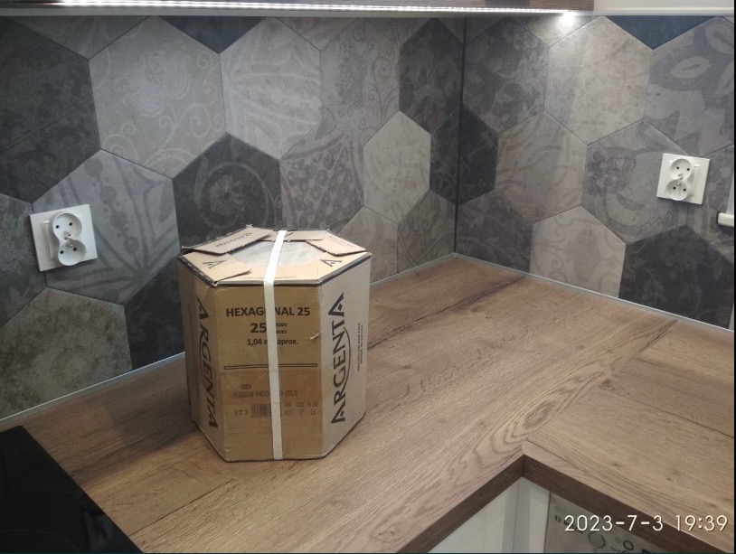 Ułożone heksagonalne płytki ścienne w odcieniach szarości i beżu, z widocznym gniazdkiem elektrycznym i kartonem płytek Argenta Hexagonal 25 na blacie imitującym drewno.