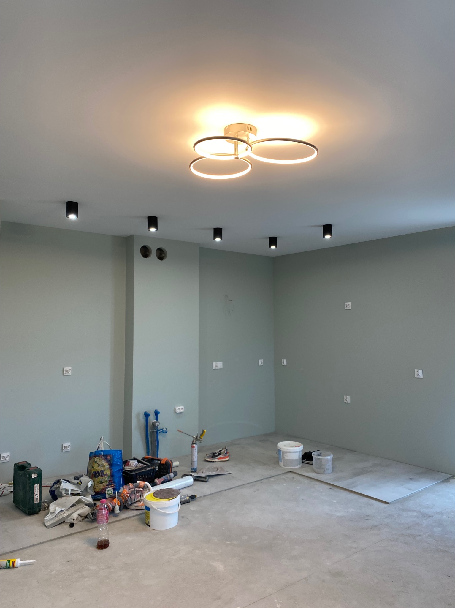 Pokój w trakcie prac, ściany pomalowane na szaro, widoczne puszki elektryczne, na podłodze narzędzia, worki, wiadra, w tle fragment ułożonych płytek, na suficie okrągła, nowoczesna lampa.