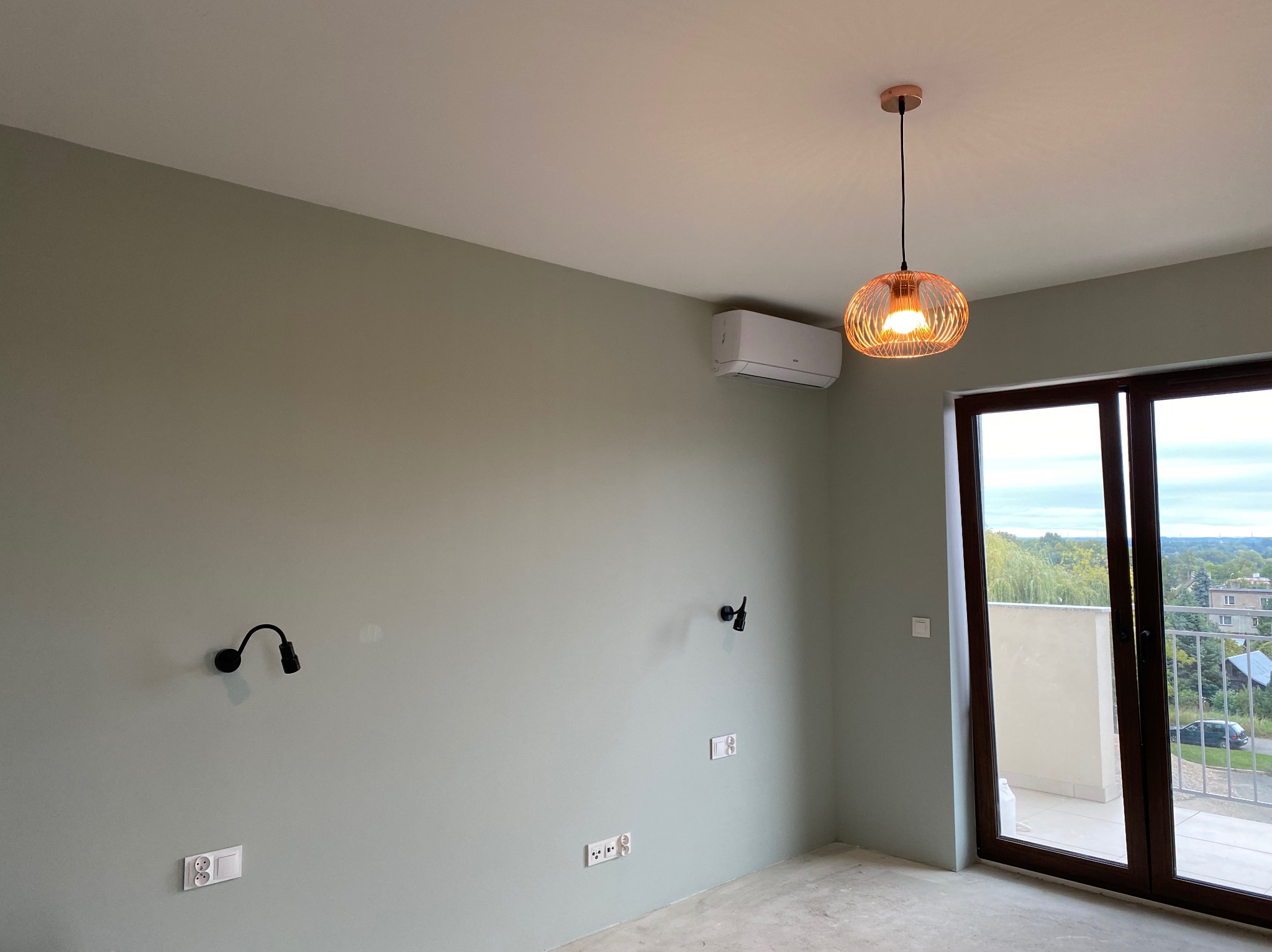 Pokój po malowaniu ścian na pistacjowy kolor, widoczny klimatyzator, ozdobna lampa wisząca, dwa kinkiety, gniazdka elektryczne i wyjście na balkon.