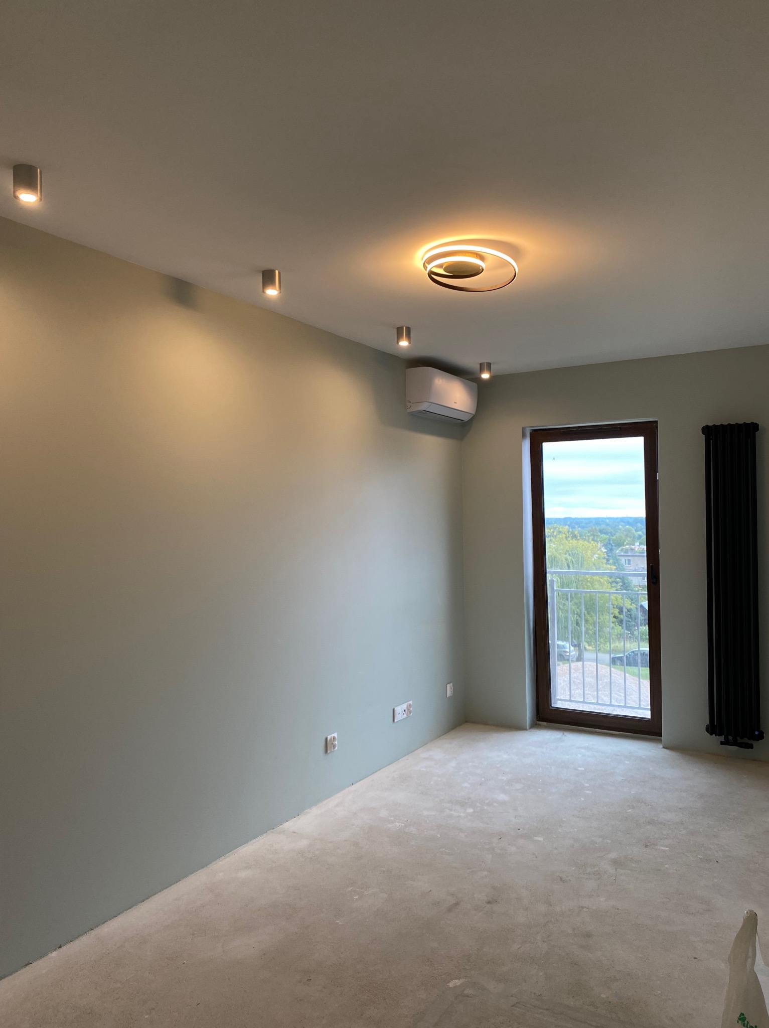 Pomalowane na jasnozielono wnętrze pokoju z zamontowanym klimatyzatorem, grzejnikiem żeberkowym, oświetleniem punktowym i nowoczesną lampą sufitową, widok na zewnątrz przez balkon.