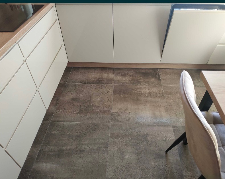 Nowoczesna kuchnia z białymi szafkami, drewnianym blatem i szarą, rustykalną podłogą z płytek. Widoczny fragment krzesła z beżową tapicerką i drewnianym stołem.