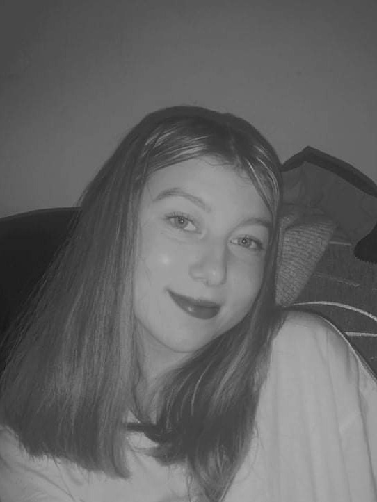 Portret młodej kobiety z długimi włosami, lekko uśmiechniętej, w czarno-białej tonacji, z delikatnym makijażem ust.