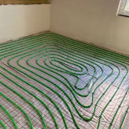 Wnętrze poddasza z ułożonym systemem ogrzewania podłogowego, widoczne zielone rury na folii z czerwoną siatką mocowane czarnymi uchwytami, surowy tynk na ścianach, drewniana konstrukcja dachu.