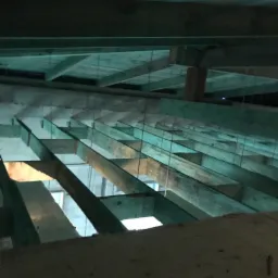 Surowy strop z drewnianych belek podwieszonych na metalowych linkach, widok z dołu na budowie, słabe oświetlenie.