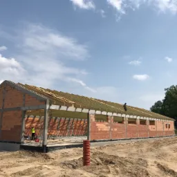 Wznoszenie ścian z czerwonej cegły i montaż drewnianej więźby dachowej na budowie hali magazynowej w słoneczny dzień.