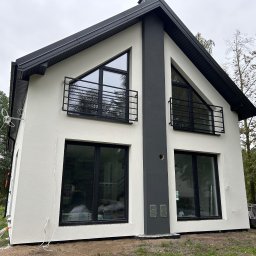 SPAW-BUD Kamil Grala - Nowoczesny dom z białą elewacją i czarnymi detalami: okna, balustrady, dach. Widoczne przewody na zewnątrz budynku. Ujęcie z boku, zieleń w tle.