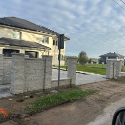 SPAW-BUD Kamil Grala - Nowoczesny dom jednorodzinny z jasną elewacją i ciemnym dachem, widoczny zza ogrodzenia z bloczków betonowych. Ułożona kostka brukowa na podjeździe.