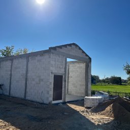 SPAW-BUD Kamil Grala - Surowy stan budynku z betonu komórkowego w słoneczny dzień. Widoczne ściany, otwór drzwiowy i okienny. Na pierwszym planie piasek i paleta z bloczkami.