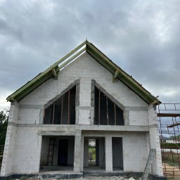 SPAW-BUD Kamil Grala - Niedokończony dom z białych bloczków, widoczne drewniane elementy dachu i rusztowanie. Surowy stan budynku pod pochmurnym niebem.