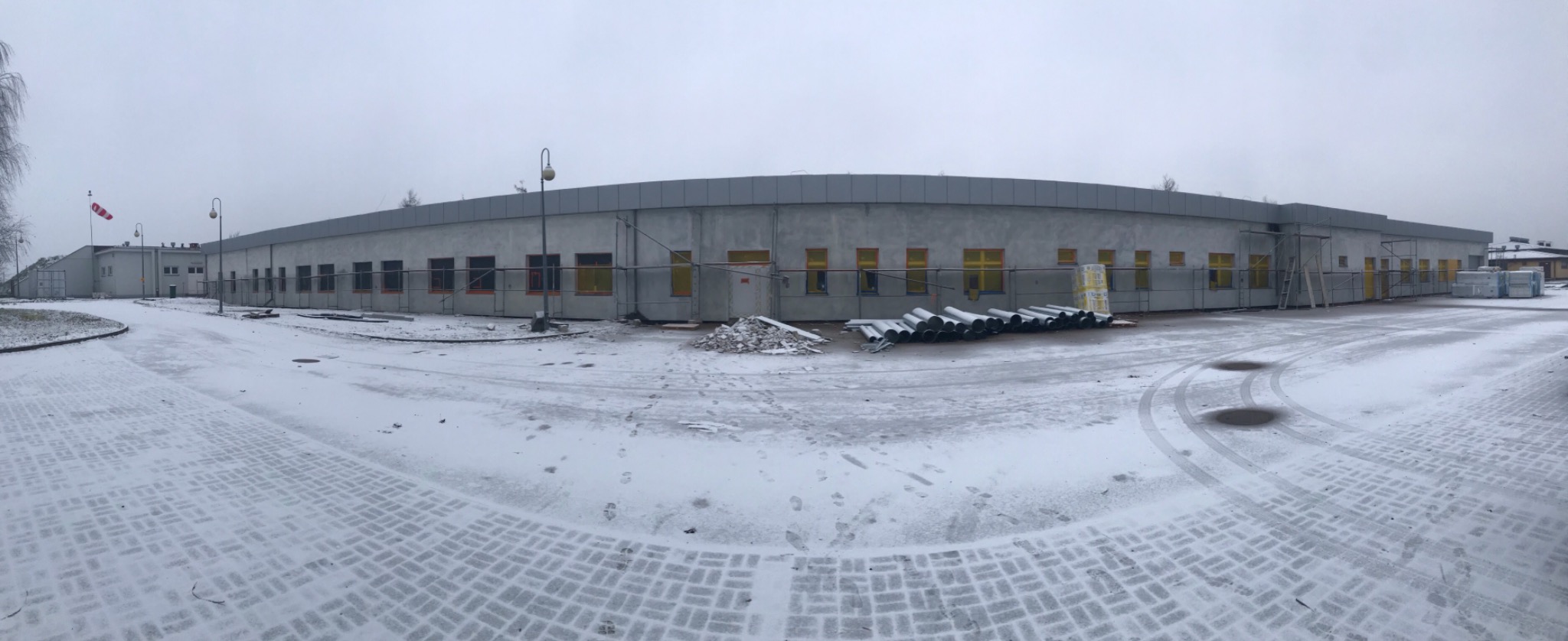 Budynek w trakcie budowy zimą, widoczne rusztowania, okna zabezpieczone folią, śnieg na ziemi i materiały budowlane.