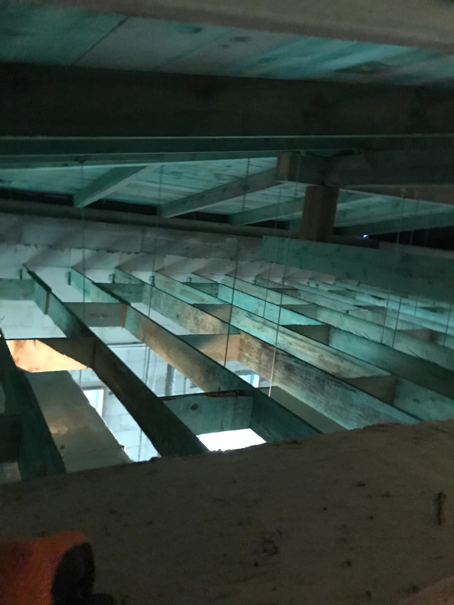 Surowy strop z drewnianych belek podwieszonych na metalowych linkach, widok z dołu na budowie, słabe oświetlenie.