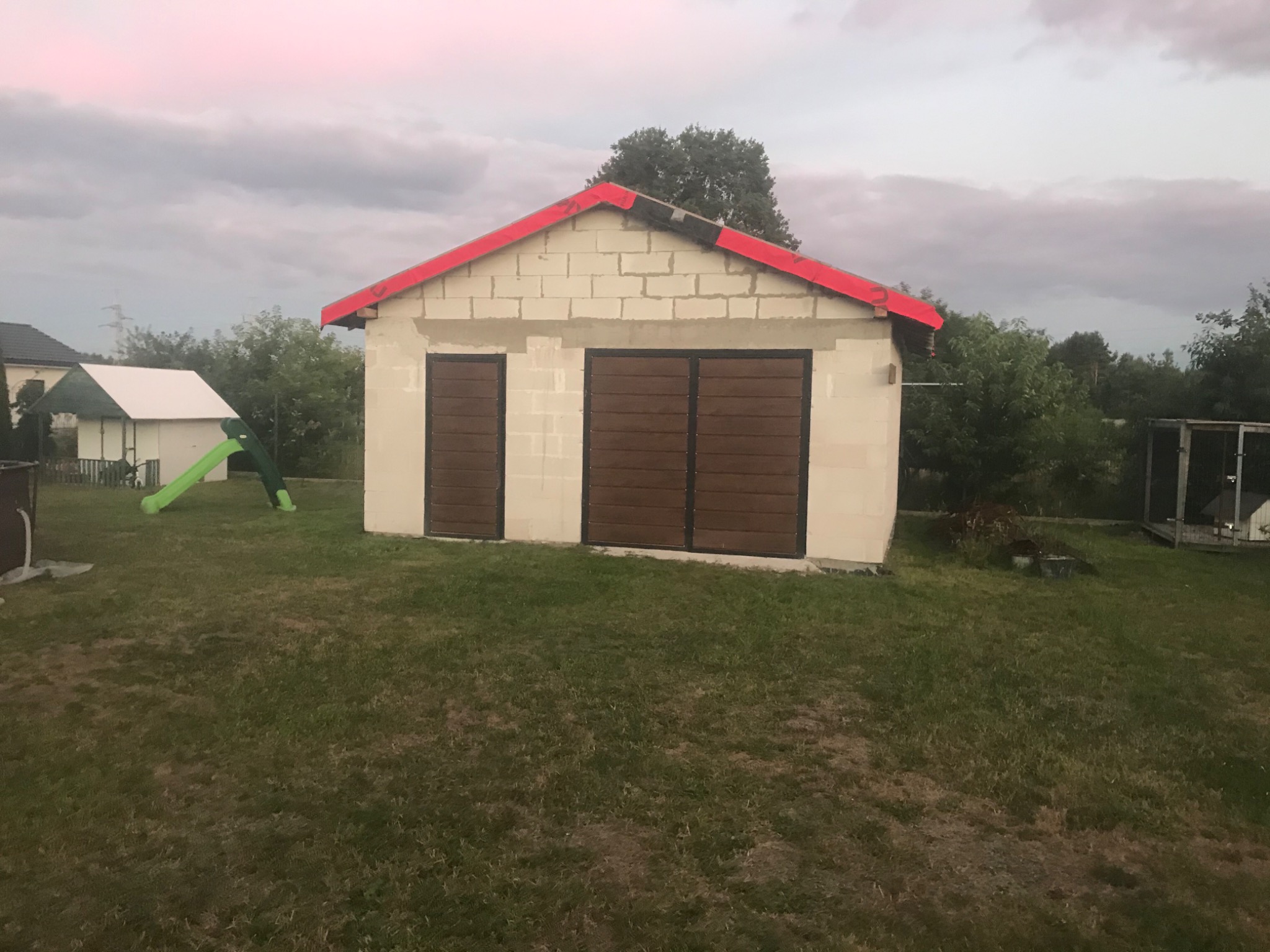 Niewielki, murowany budynek gospodarczy z czerwonym dachem w Ostrołęce, z dwoma brązowymi bramami garażowymi, w trakcie budowy, widoczny na tle trawnika i innych budynków.