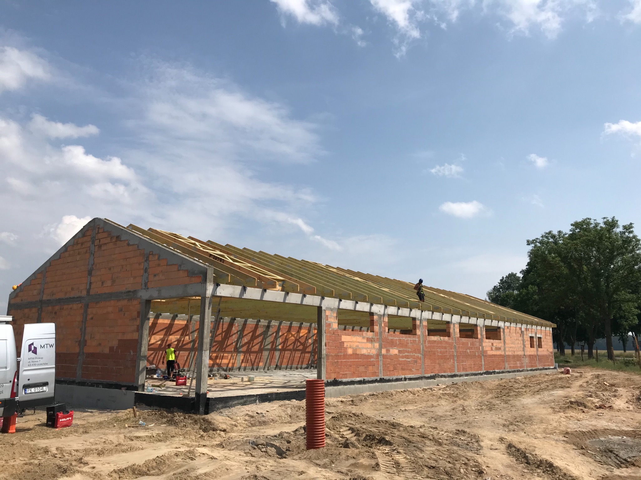 Wznoszenie ścian z czerwonej cegły i montaż drewnianej więźby dachowej na budowie hali magazynowej w słoneczny dzień.