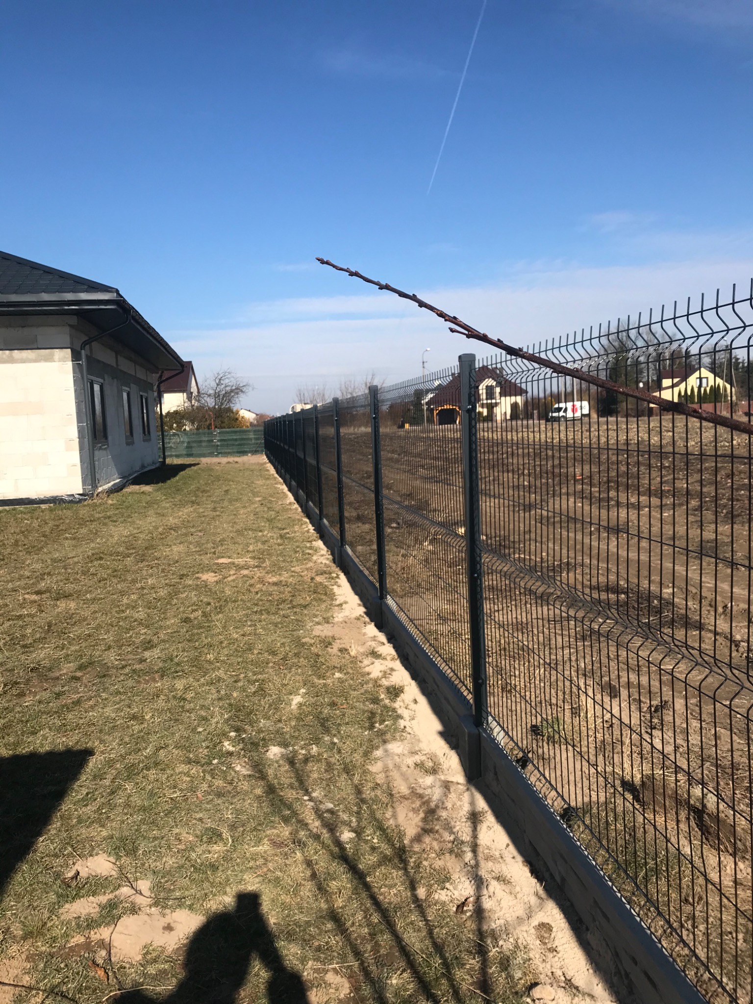 Nowoczesne ogrodzenie panelowe w kolorze grafitowym, oddzielające posesję z budynkiem w stanie surowym od niezagospodarowanego terenu z widocznymi domami w tle.