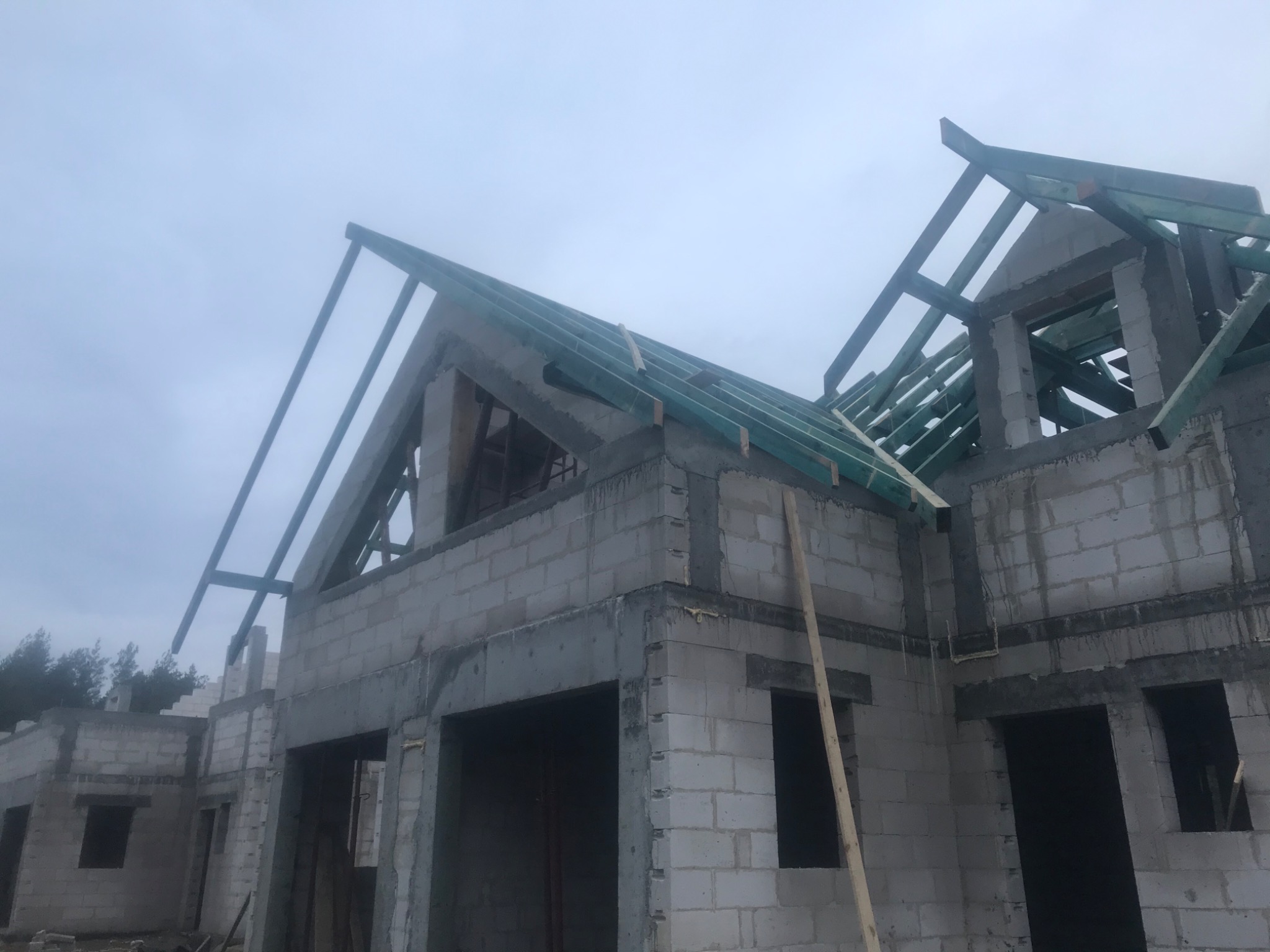 Surowy stan domu z betonu komórkowego, widoczne ściany i konstrukcja dachu z zielonymi elementami, brak okien i drzwi, pochmurne niebo w tle.