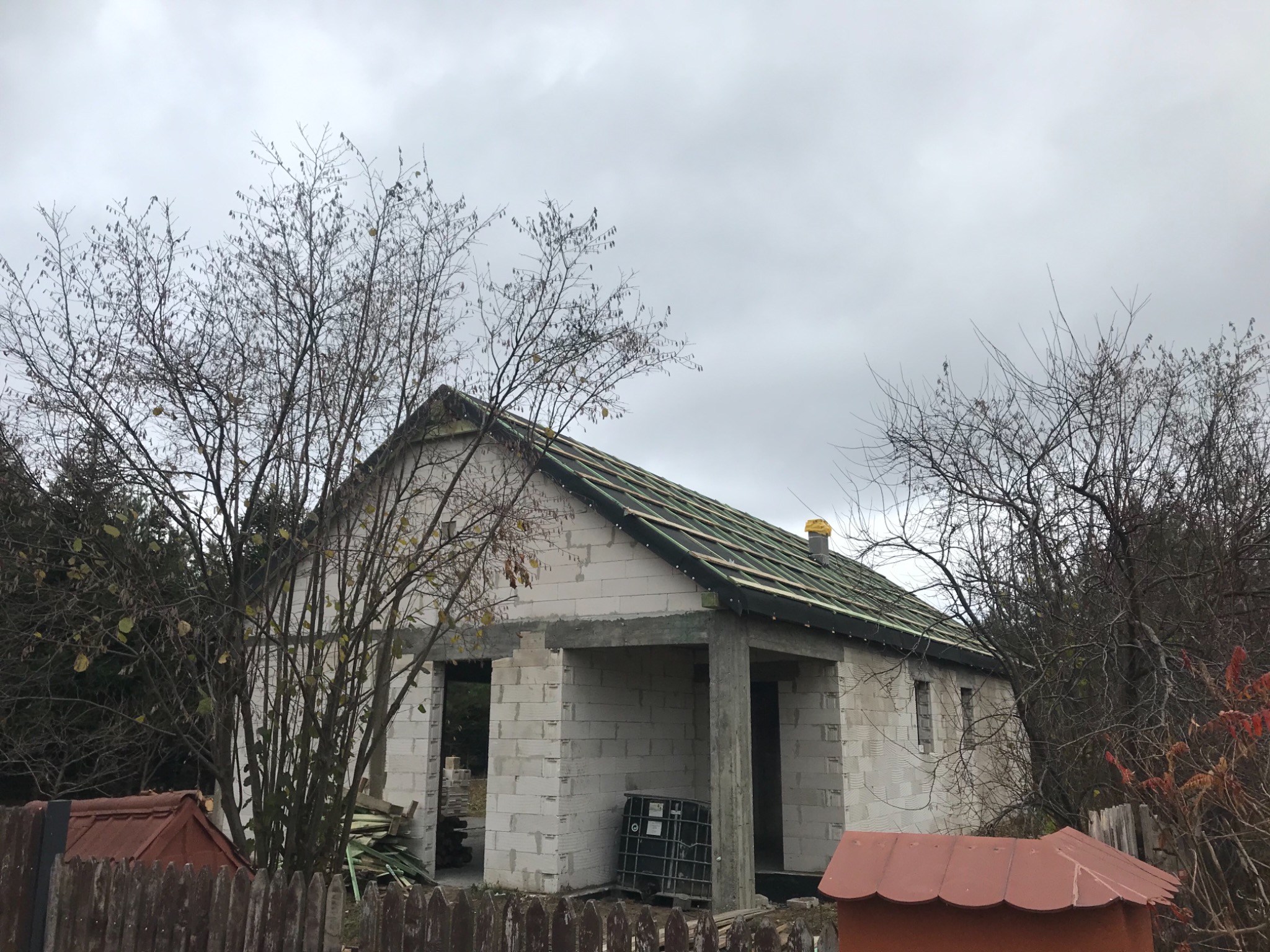 Niedokończony dom murowany z betonu komórkowego, widoczne drewniane łaty dachowe i niezadaszona weranda, otoczony drzewami bez liści pod pochmurnym niebem.