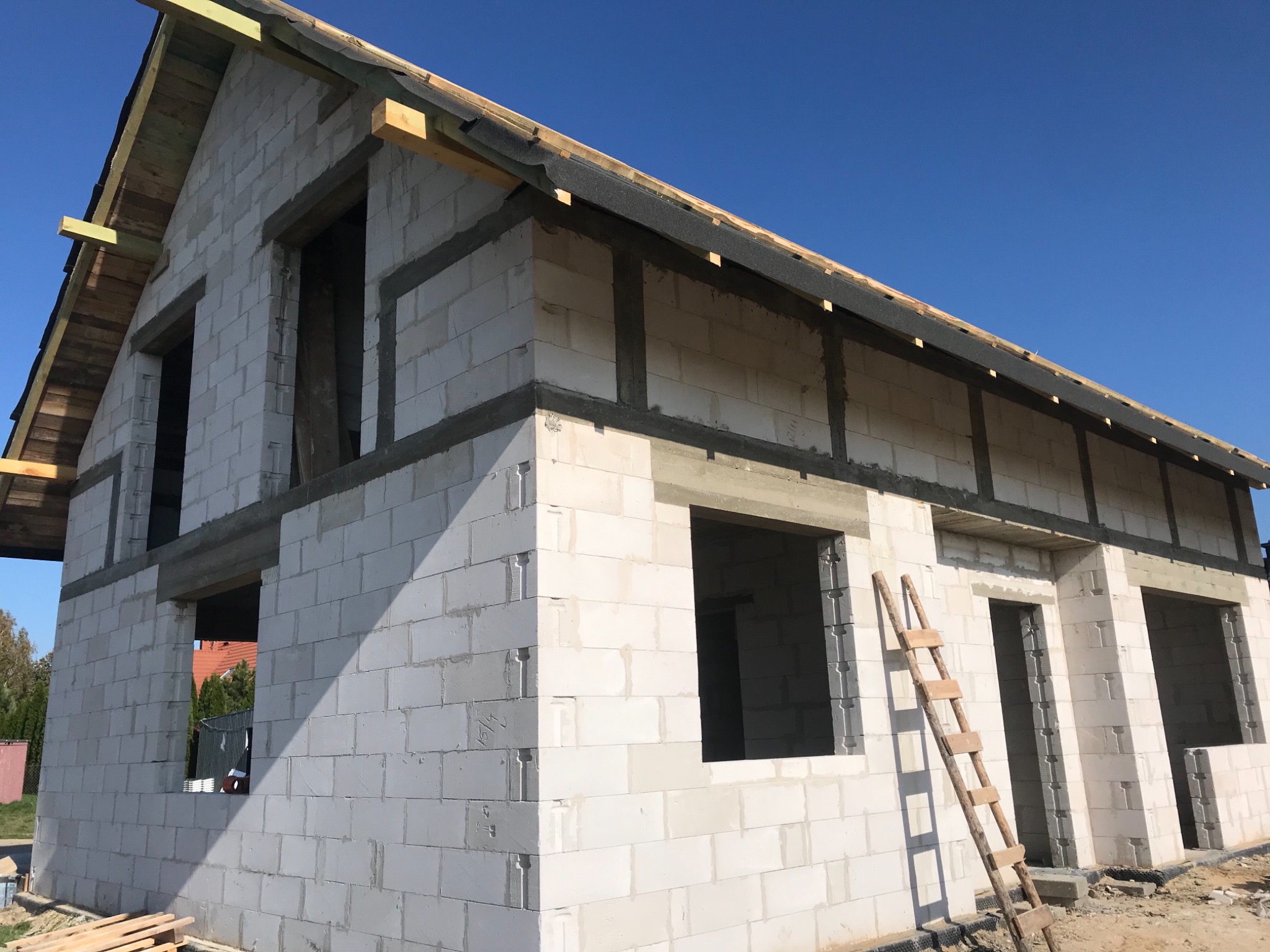 Surowy stan domu z białego pustaka, z drewnianą konstrukcją dachu i drabiną opartą o ścianę.