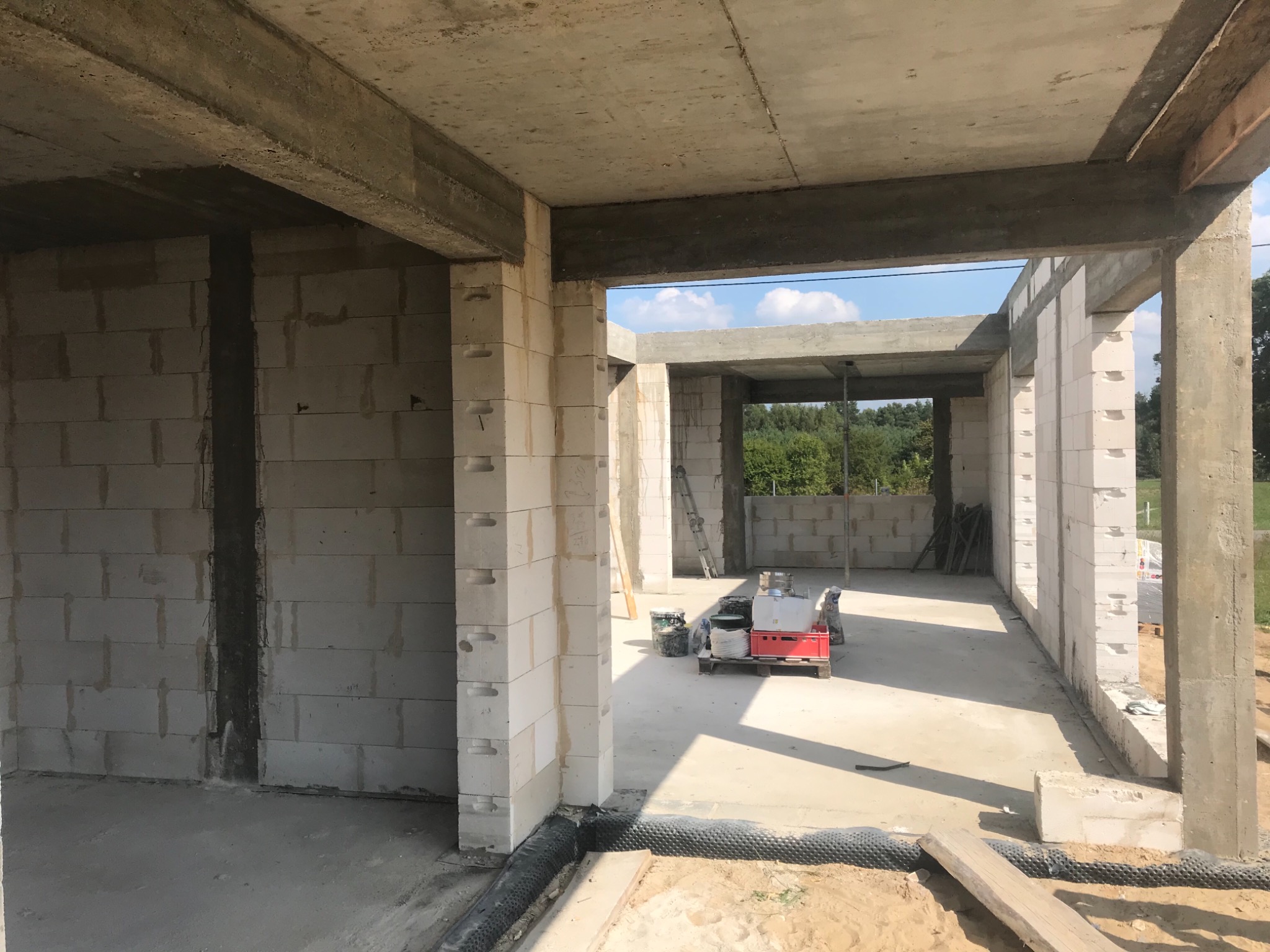 Surowy stan domu z pustaków, widoczne ściany, betonowe nadproża, w tle okno z widokiem na zieleń, na podłodze materiały budowlane na palecie.