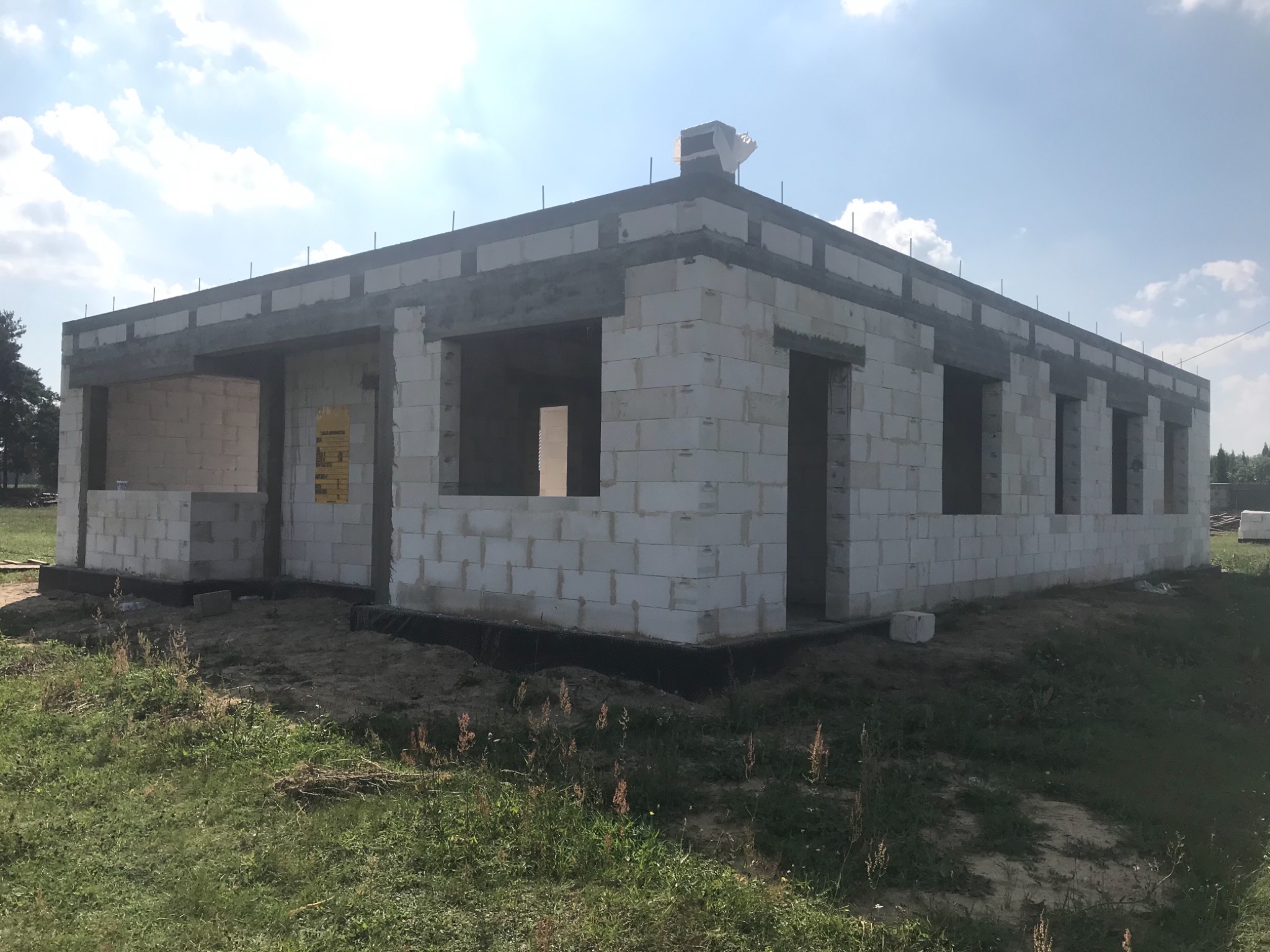 Budowa domu jednorodzinnego z białych bloczków, widoczne zbrojenie w wieńcu, etap surowy otwarty, teren wokół niezagospodarowany, w tle drzewa.