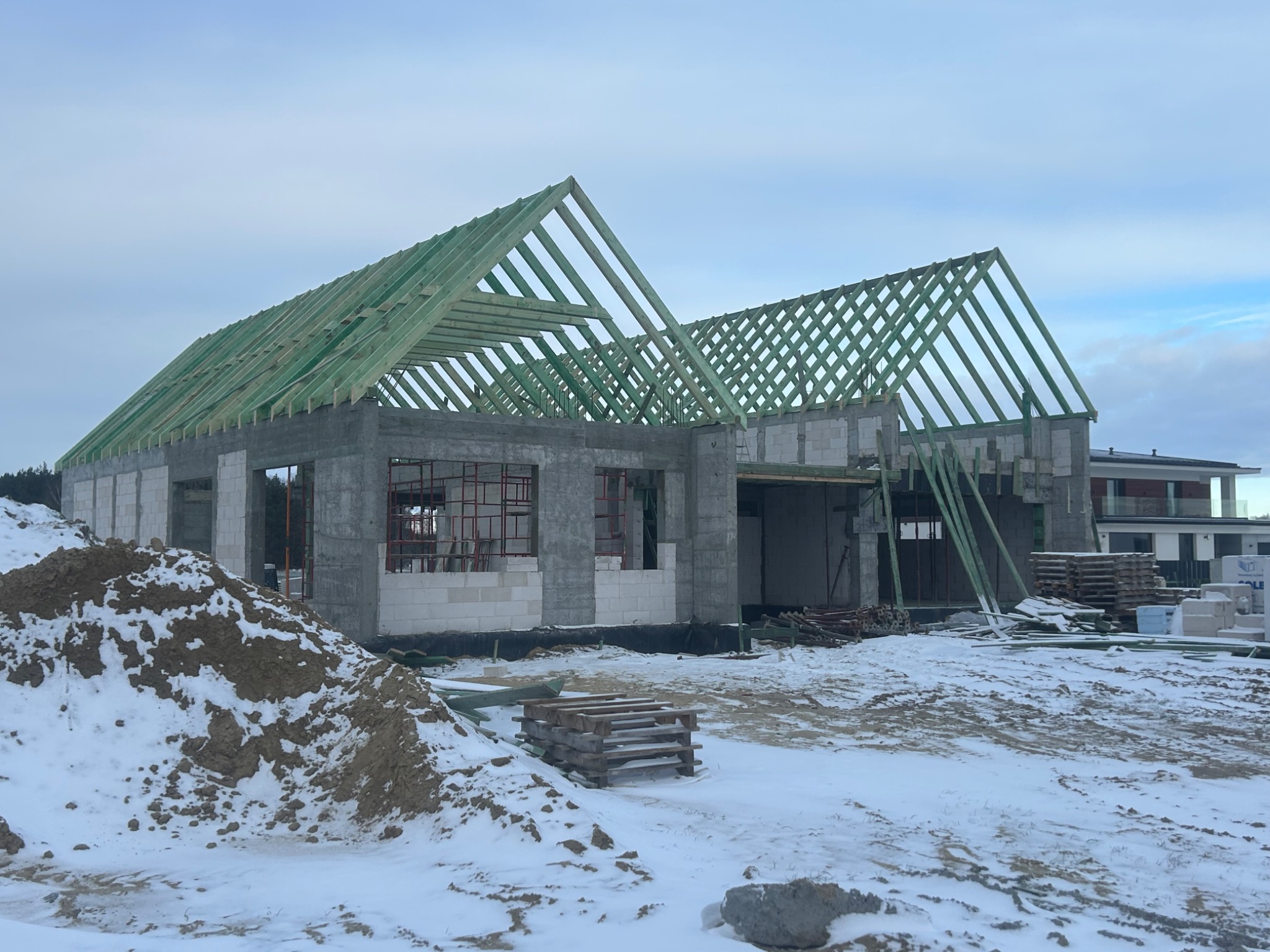 Surowy stan domu z betonu komórkowego w zimowej scenerii. Widoczne ściany, zielona konstrukcja dachu i zaspy śniegu. Palety i gruz budowlany na pierwszym planie.