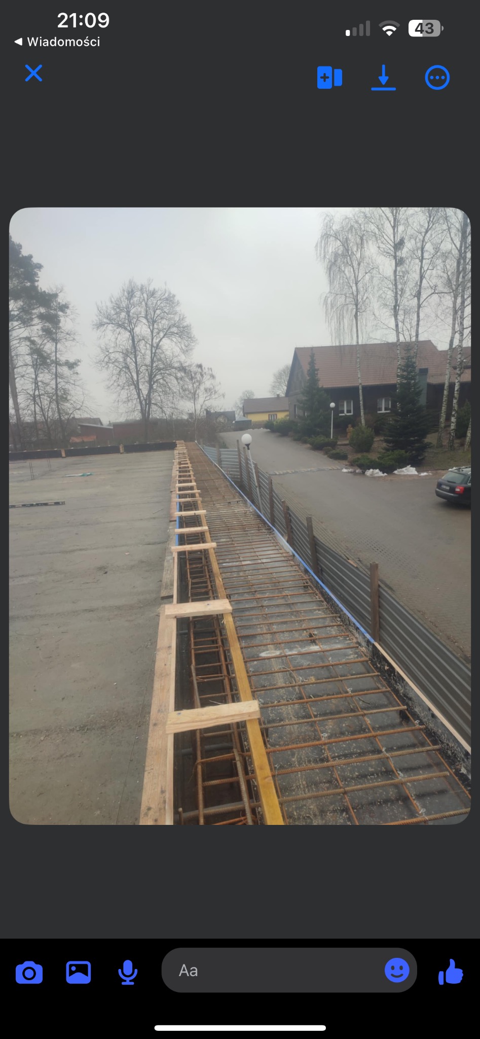 Zbrojenie fundamentów z drewnianym szalunkiem na placu budowy w pochmurny dzień. Widoczne drzewa w tle i fragment zabudowań. Ujęcie z góry.