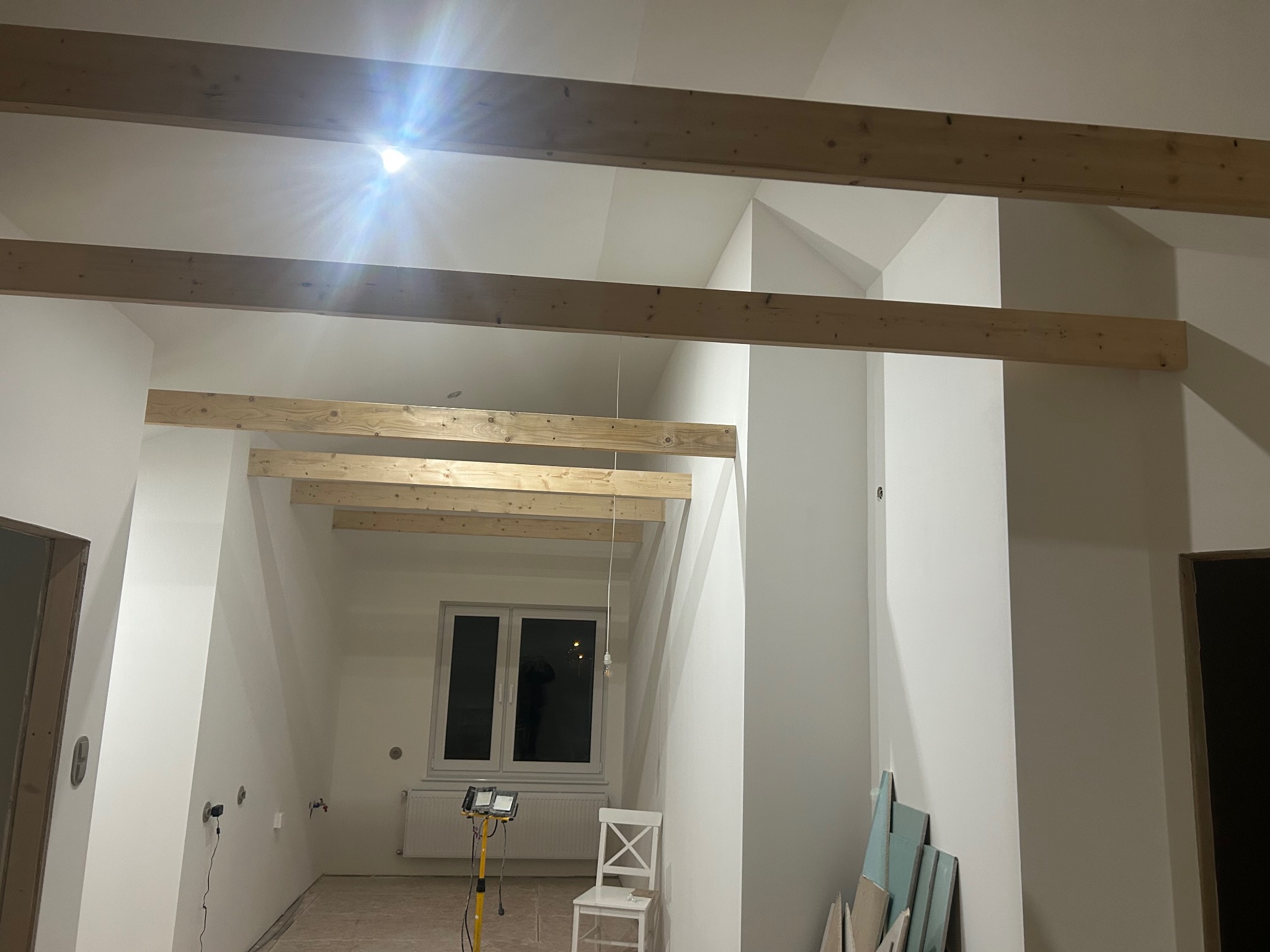 Wnętrze poddasza w trakcie prac wykończeniowych. Widoczne drewniane belki stropowe, białe ściany, okno i lampa budowlana. Częściowo położona podłoga.