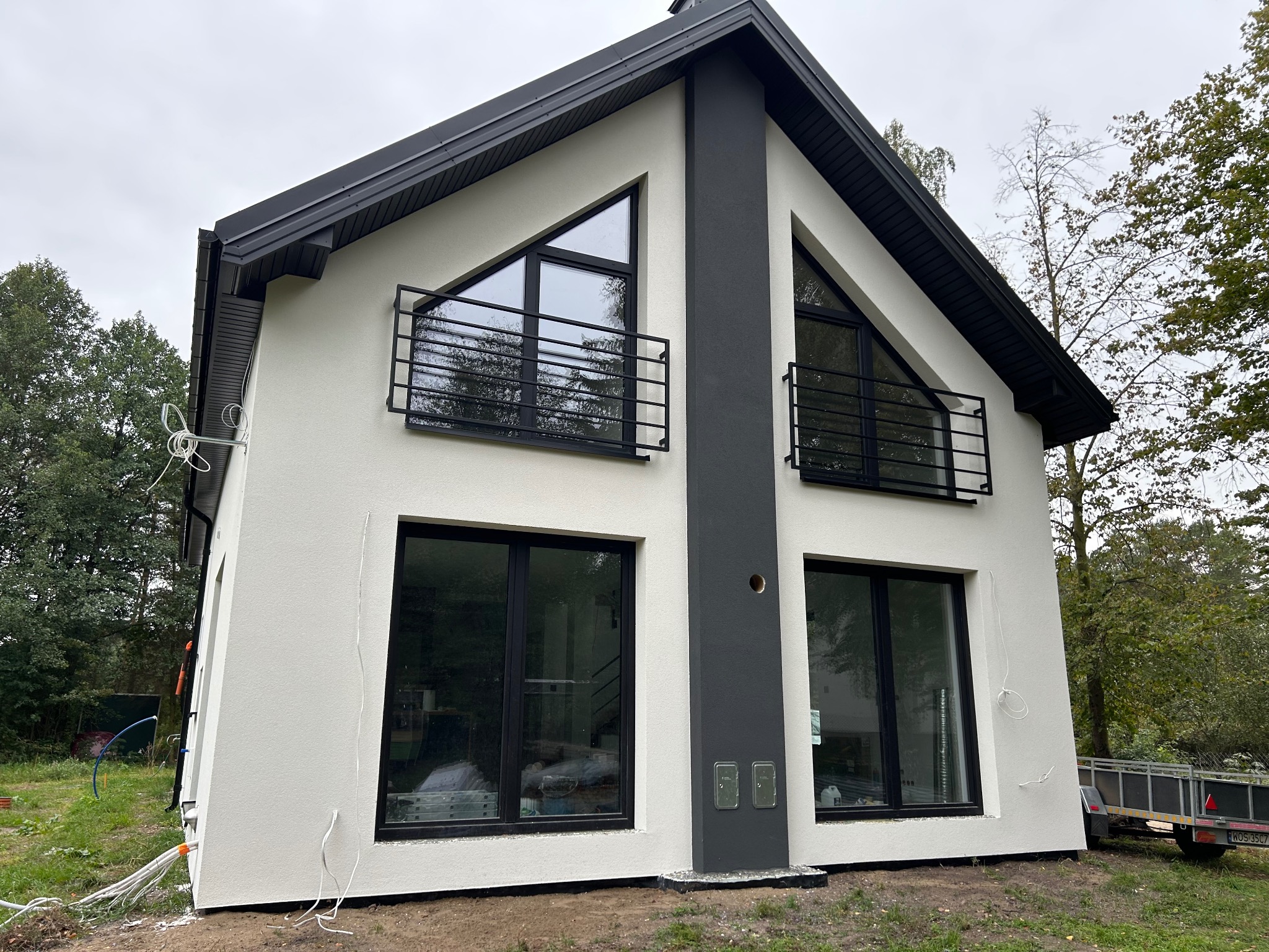 Nowoczesny dom z białą elewacją i czarnymi detalami: okna, balustrady, dach. Widoczne przewody na zewnątrz budynku. Ujęcie z boku, zieleń w tle.
