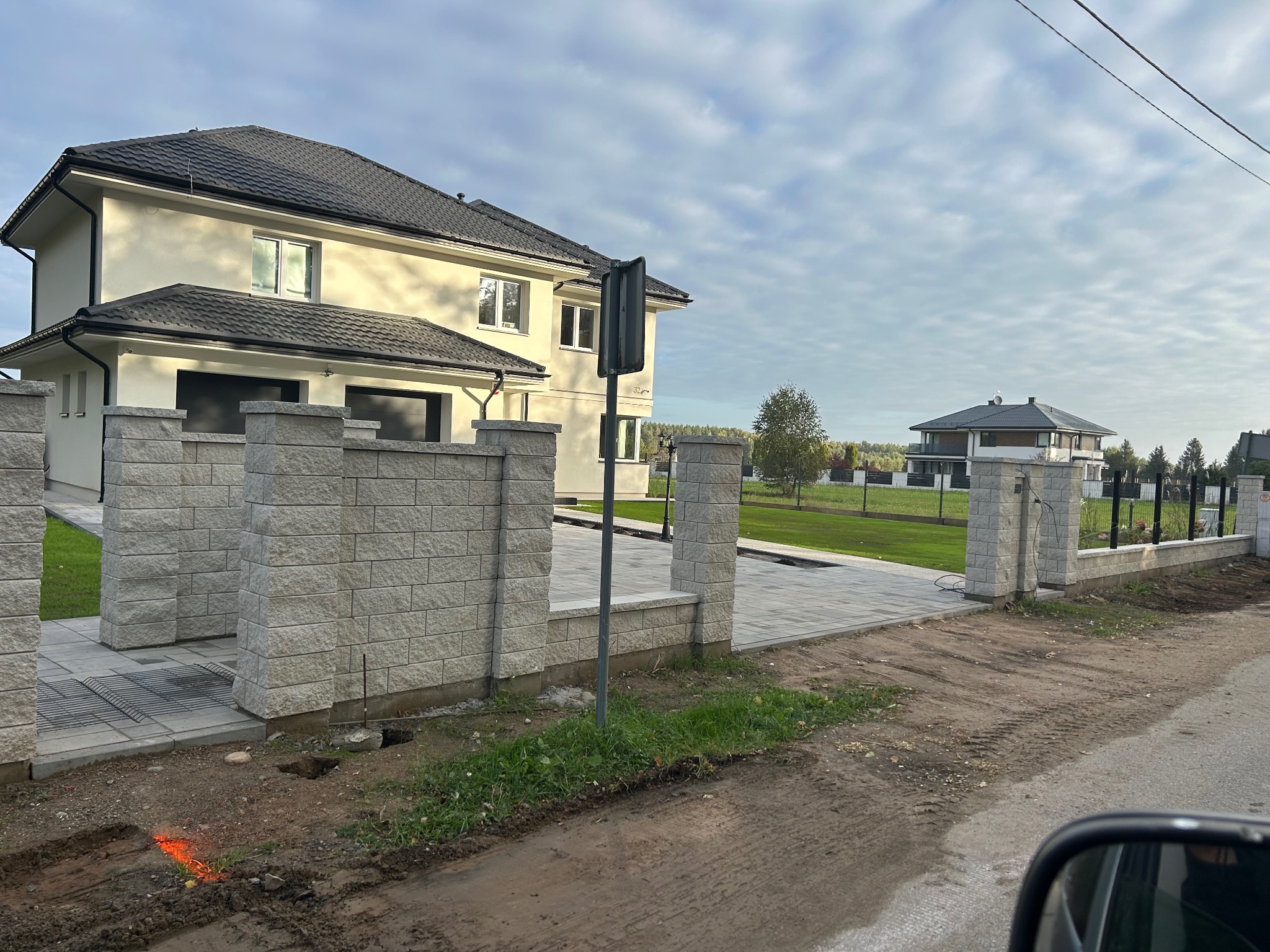 Nowoczesny dom jednorodzinny z jasną elewacją i ciemnym dachem, widoczny zza ogrodzenia z bloczków betonowych. Ułożona kostka brukowa na podjeździe.