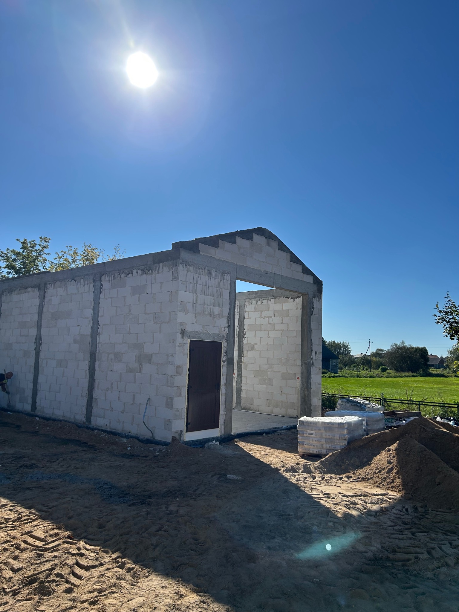 Surowy stan budynku z betonu komórkowego w słoneczny dzień. Widoczne ściany, otwór drzwiowy i okienny. Na pierwszym planie piasek i paleta z bloczkami.
