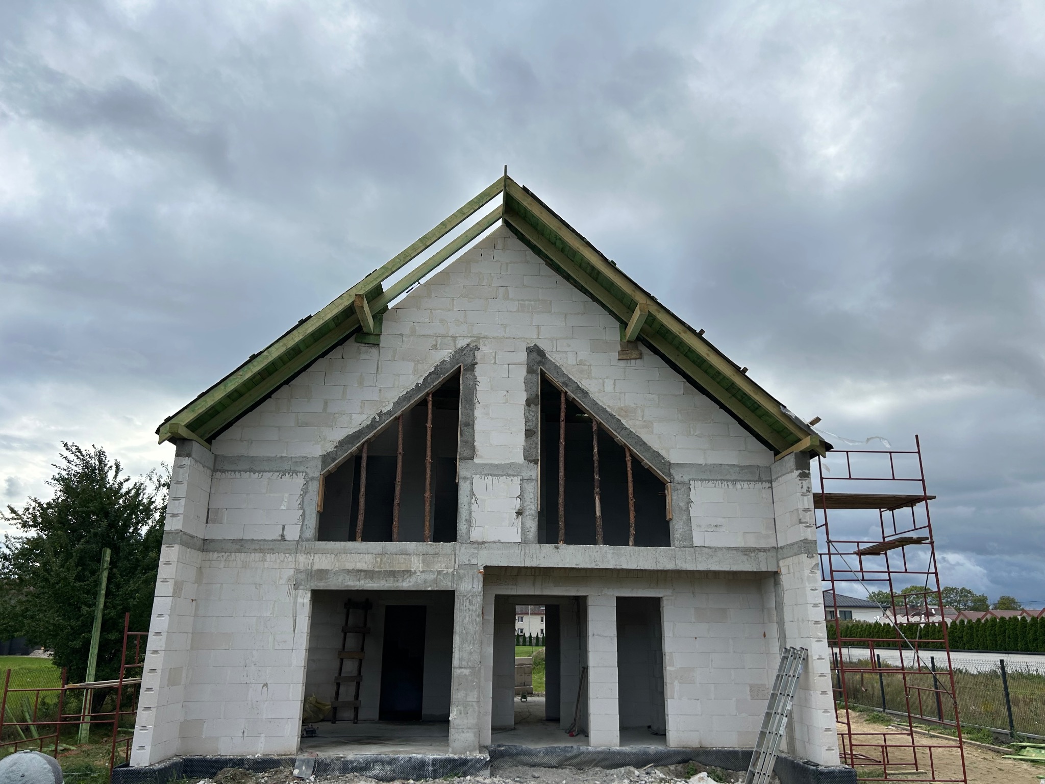 Niedokończony dom z białych bloczków, widoczne drewniane elementy dachu i rusztowanie. Surowy stan budynku pod pochmurnym niebem.