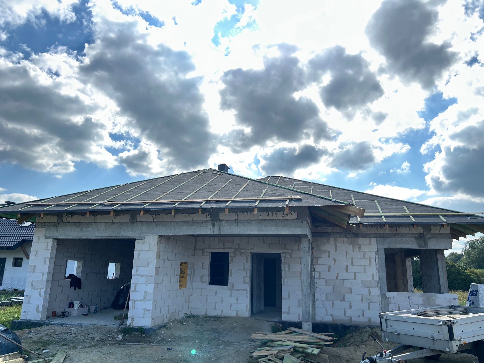 Niedokończony dom z białego pustaka pod ciemnym dachem na tle pochmurnego nieba. Widoczne drewniane elementy konstrukcji dachu i porozrzucane deski na ziemi.