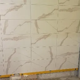 Ściana w trakcie układania płytek imitujących marmur, widoczne krzyżyki dystansowe i poziomica, fragment ściany przygotowany pod płytki, obok wystające rurki instalacji wodnej.