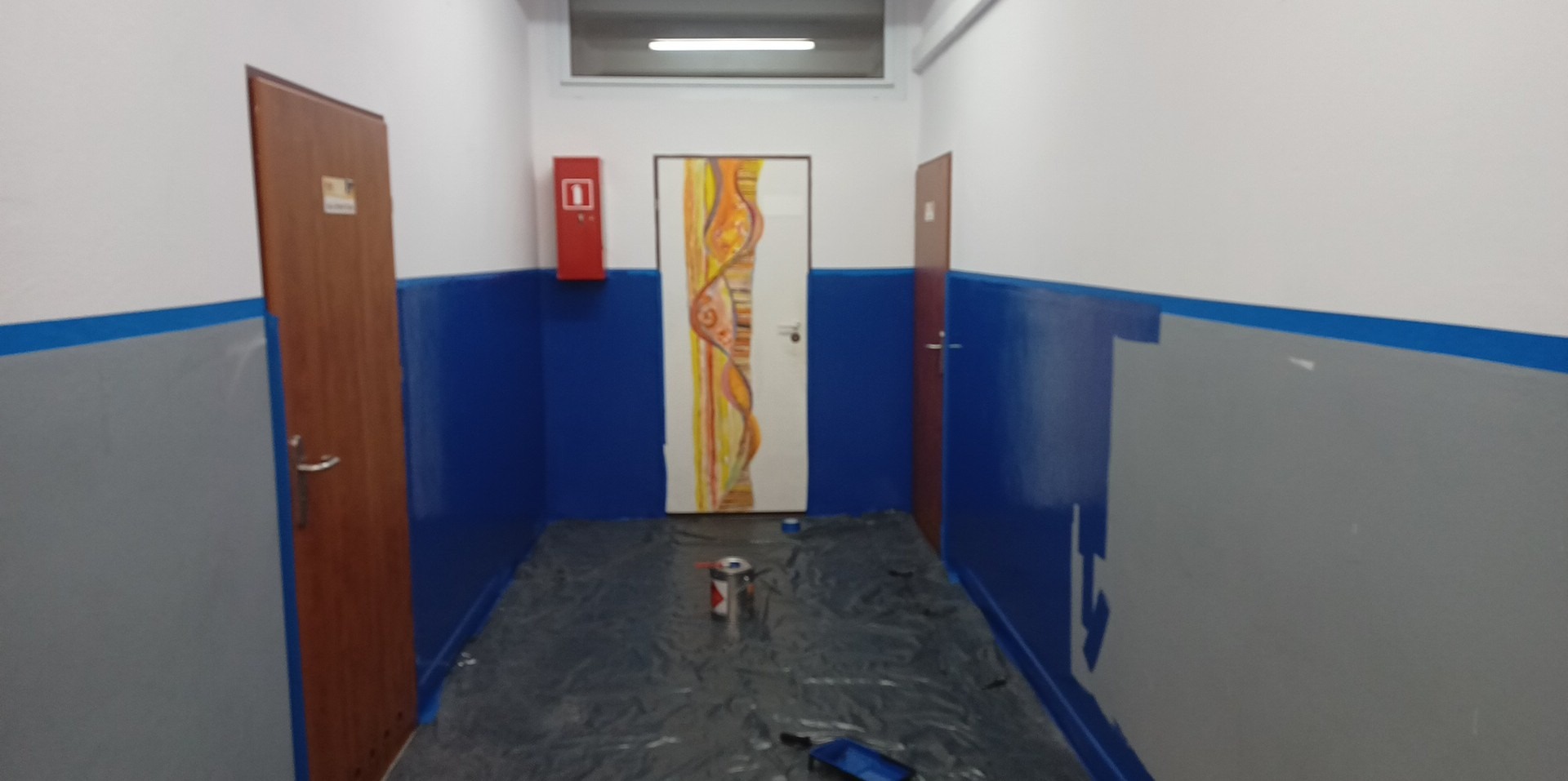 Korytarz w trakcie malowania na niebiesko i szaro, zabezpieczony folią, widoczna puszka z farbą i kuweta malarska, drzwi z abstrakcyjnym malunkiem.