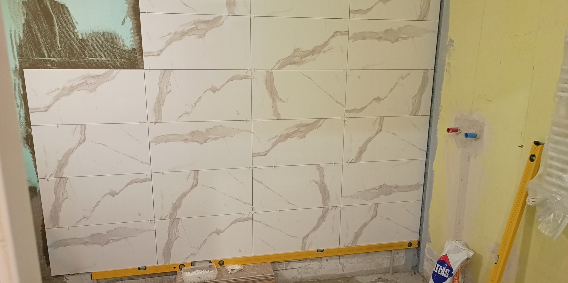 Ściana w trakcie układania płytek imitujących marmur, widoczne krzyżyki dystansowe i poziomica, fragment ściany przygotowany pod płytki, obok wystające rurki instalacji wodnej.