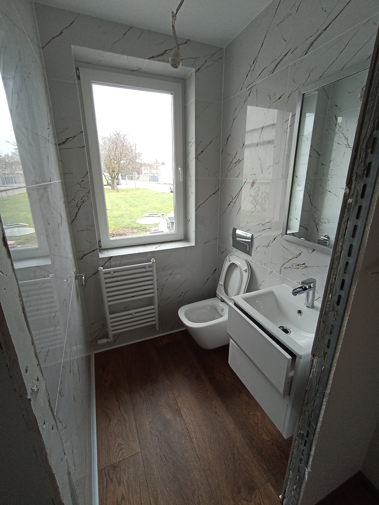 Ukończona łazienka z płytkami imitującymi marmur, drewnianą podłogą, białą umywalką z szafką i grzejnikiem drabinkowym, widok z perspektywy podłogi, z fragmentem niezakończonej zabudowy po prawej...