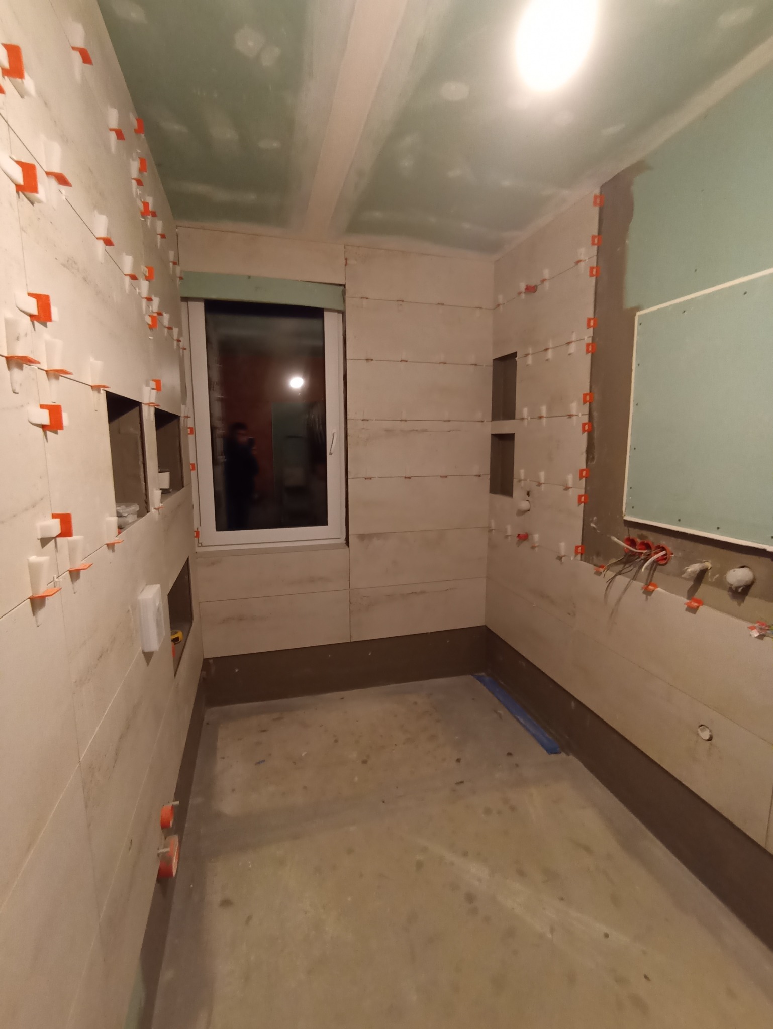Wnętrze łazienki w trakcie układania płytek ściennych, widoczne poziomowanie z użyciem krzyżyków dystansowych, przygotowana podłoga i częściowo zabudowana ściana z płyty gipsowo-kartonowej, okno...