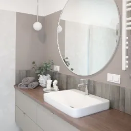 Nowoczesna łazienka w stonowanych barwach: beż, biel i szarość. Umywalka nablatowa, okrągłe lustro, dekoracyjne ręczniki i elementy dekoracyjne. Pralka zabudowana pod blatem.