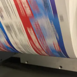 Drukarka rotacyjna w trakcie drukowania arkusza z pasami w kolorach czerwonym, białym i niebieskim, widoczne cienkie szare linie.