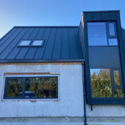 Nowoczesny dom w trakcie budowy z ciemnoszarym dachem blaszanym, dwoma oknami dachowymi i niestandardową obudową okienną, widoczne niedokończone elewacje.