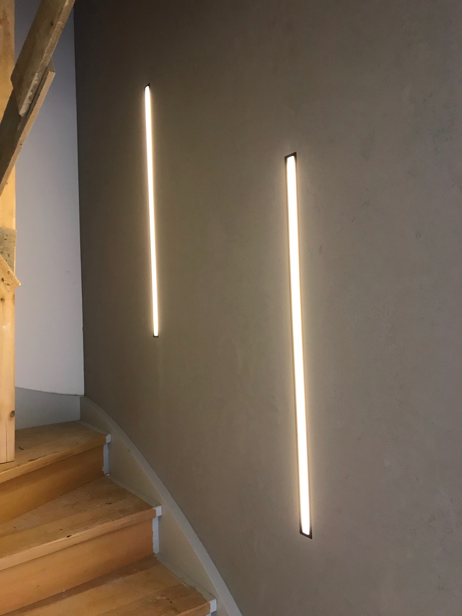 Klatka schodowa z drewnianymi schodami i dwiema pionowymi lampami LED wbudowanymi w szarą ścianę, widoczny fragment drewnianej konstrukcji schodów.