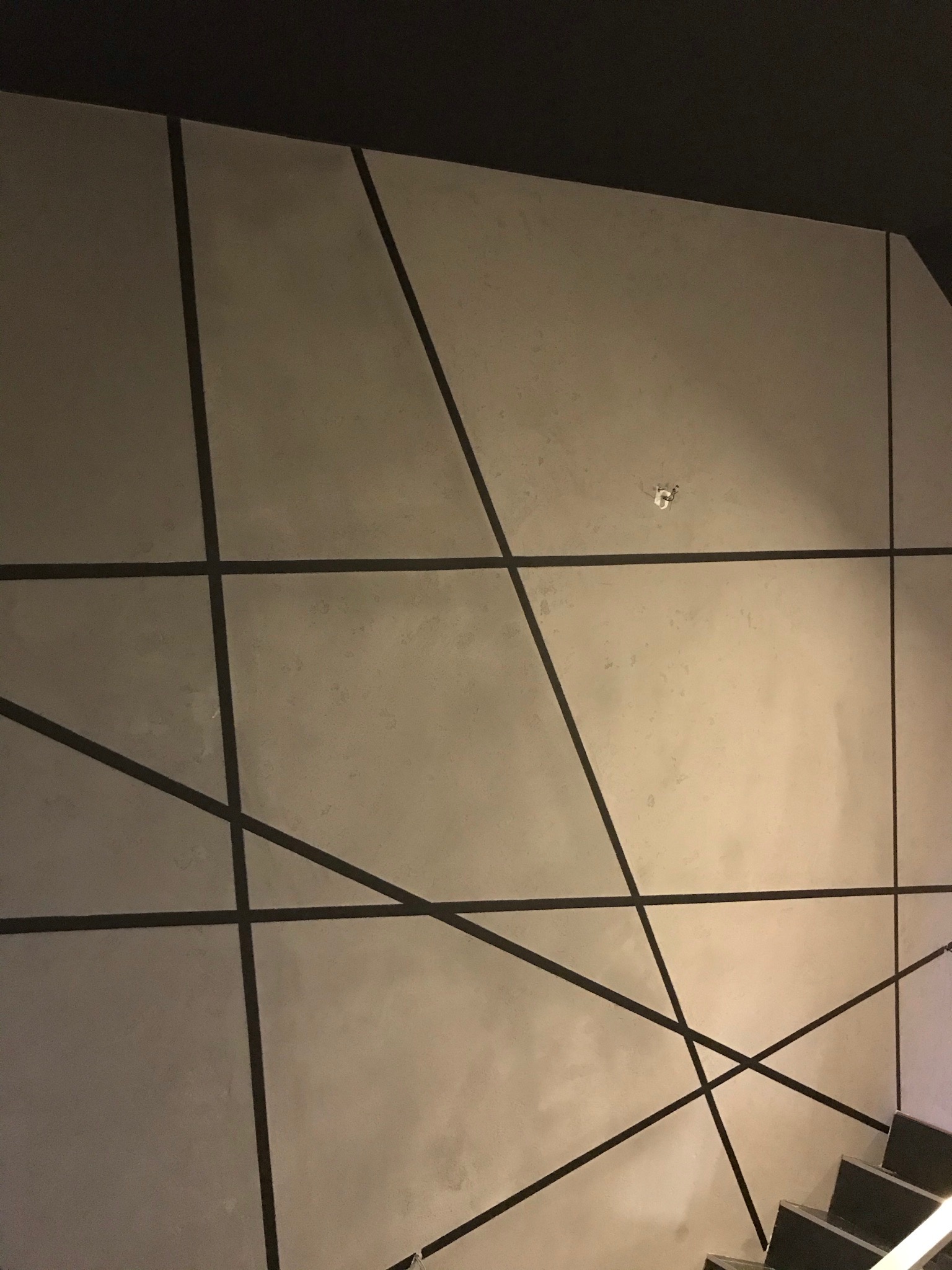 Ściana z szarego tynku dekoracyjnego z czarnymi, geometrycznymi liniami, widziana z dołu przy schodach.