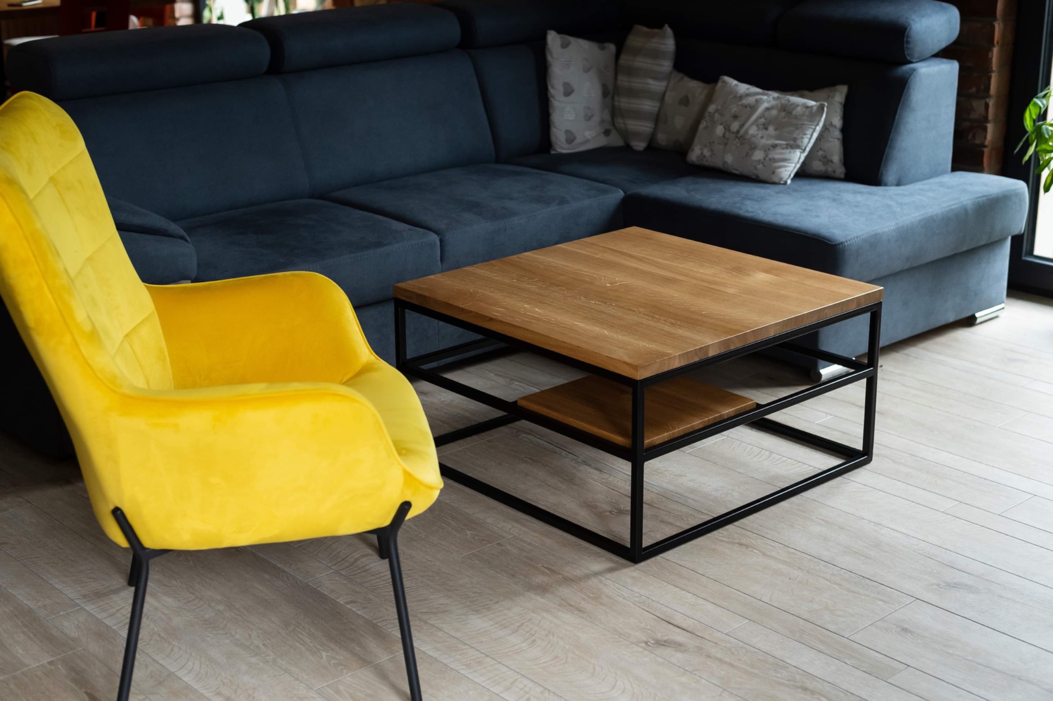Wnętrze salonu z drewnianą podłogą, narożną sofą w odcieniach granatu, żółtym welurowym fotelem i stolikiem kawowym z drewnianym blatem na metalowej konstrukcji.