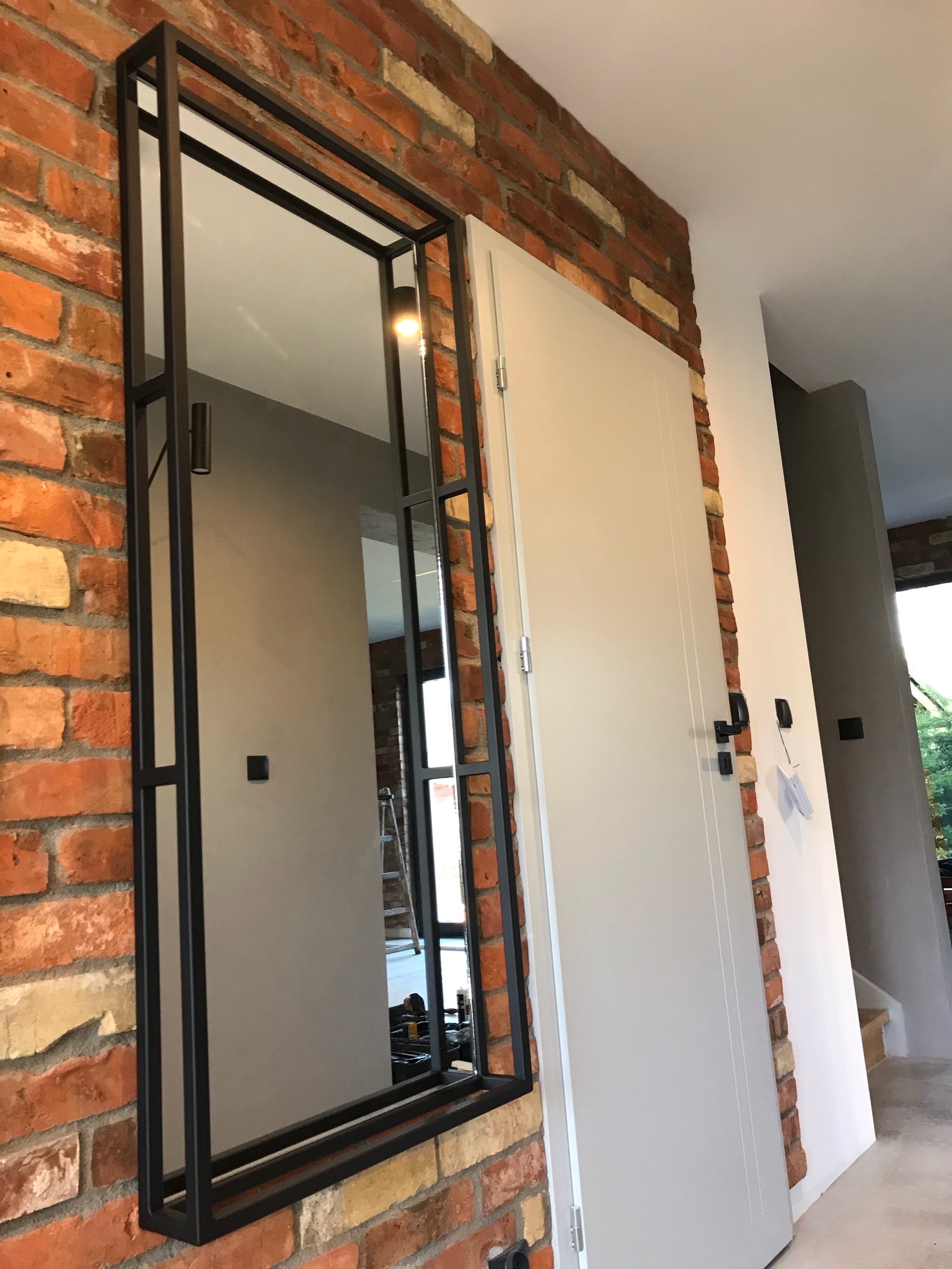 Wysokie lustro w czarnej, metalowej ramie zawieszone na ścianie z czerwonej cegły, obok białe drzwi i fragment klatki schodowej w tle.