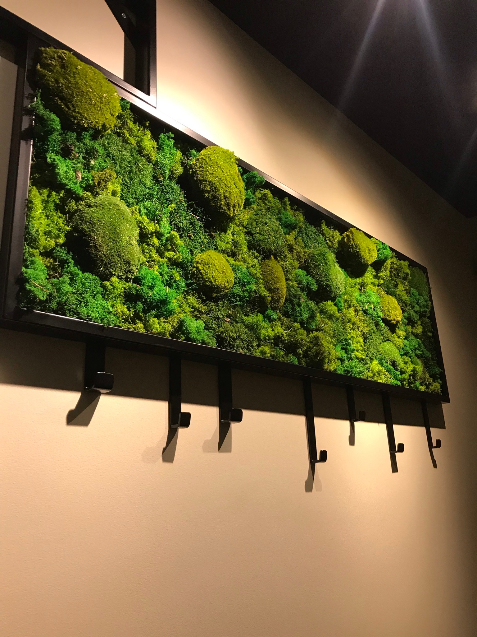 Panel z mchu stabilizowanego w czarnej ramie z haczykami, wiszący na beżowej ścianie, element dekoracyjny wnętrza.