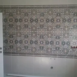 Ściana w łazience wyłożona dekoracyjnymi płytkami ceramicznymi w odcieniach beżu, szarości i granatu, z widoczną białą ramą drzwi po lewej stronie i wystającymi rurami instalacji wodnej na środku...